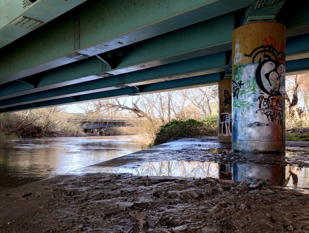 Photo: graffiti on pillars, and muddy puddles, under a bridge beside a river.