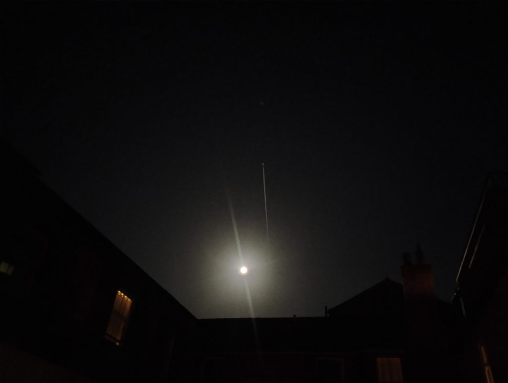 Photo: a blurry moon in a black sky, with an aeroplane's contrail highlighted by its glare.