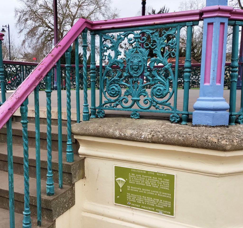 Photo: part of a park bandstand, with railings painted pink, blue, and teal. A green plaque tells the story of a Mr John Hampton, "the first person to make a successful descent by a parachute" on October 3rd, 1838, "from a balloon which ascended from these gardens".