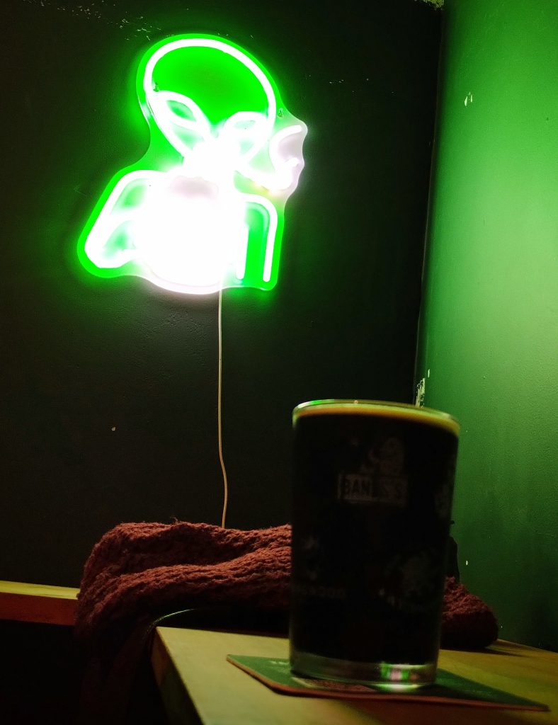 Phot of a small glass of beer in front of a neon light shaped like an alien.