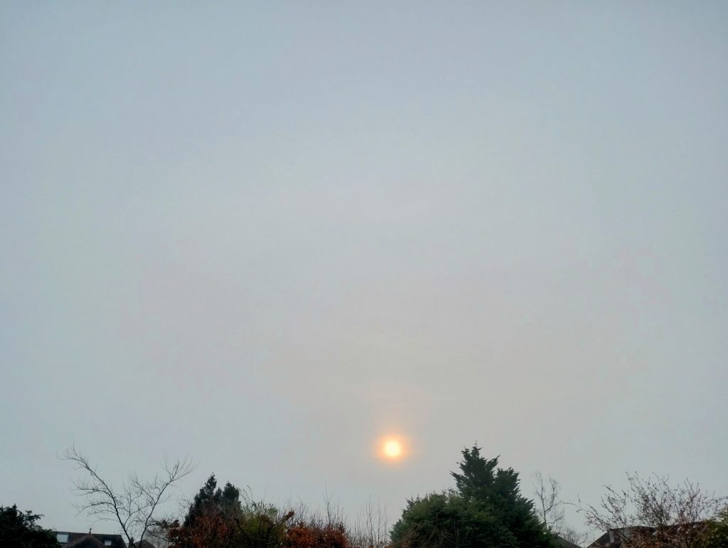 Photo: hazy grey sky with orange sun shining dimly through like a torch.