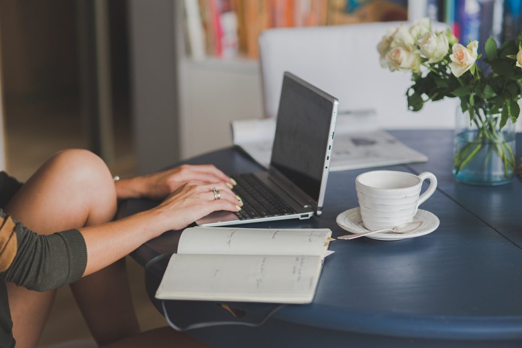 Self-editing a novel: Photo of a woman's hands at a laptop on a round blue table, with a notebook, white mug and teaspoon, and vase of roses nearby.
